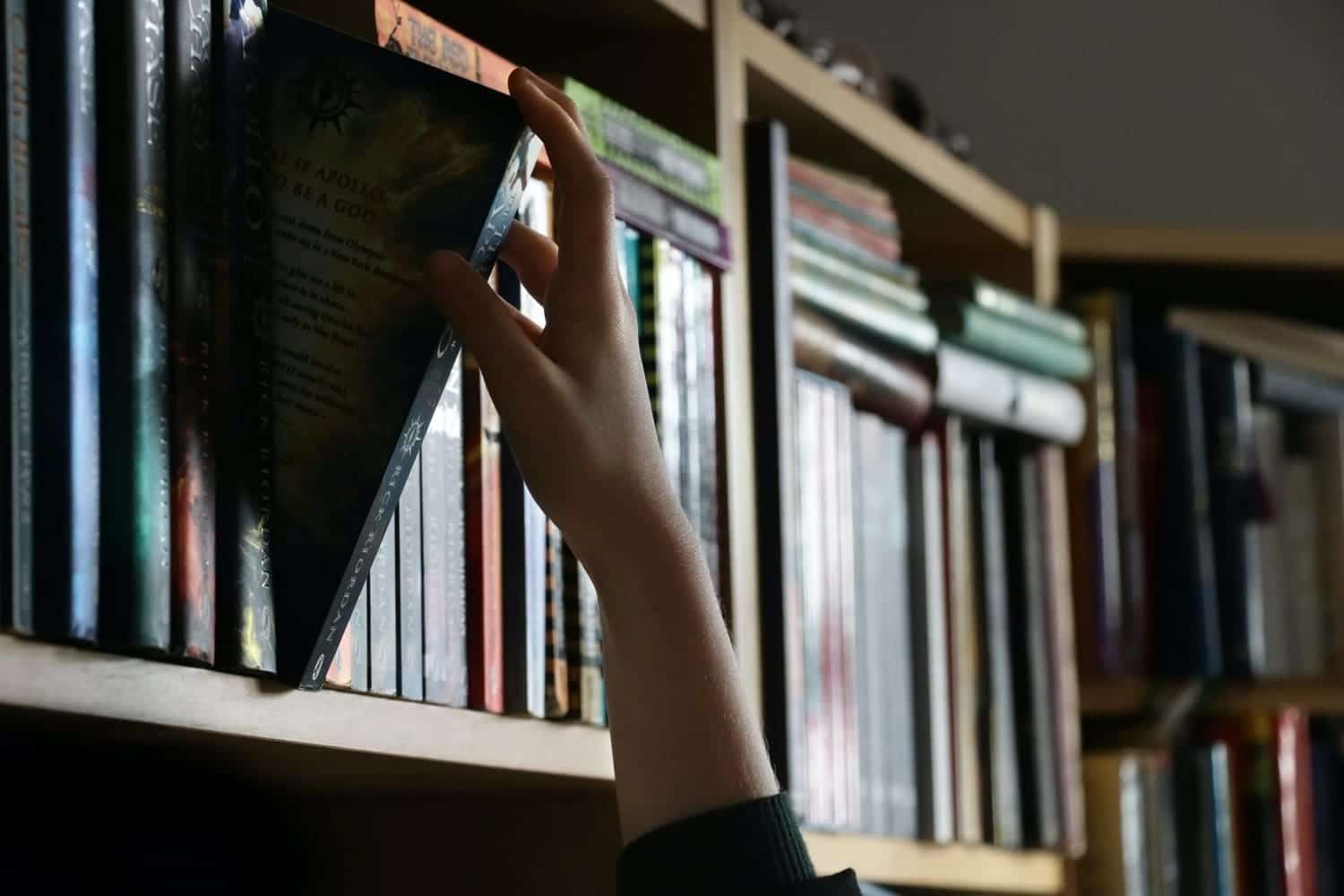 A person's hand picking a book from a shelf in a library.